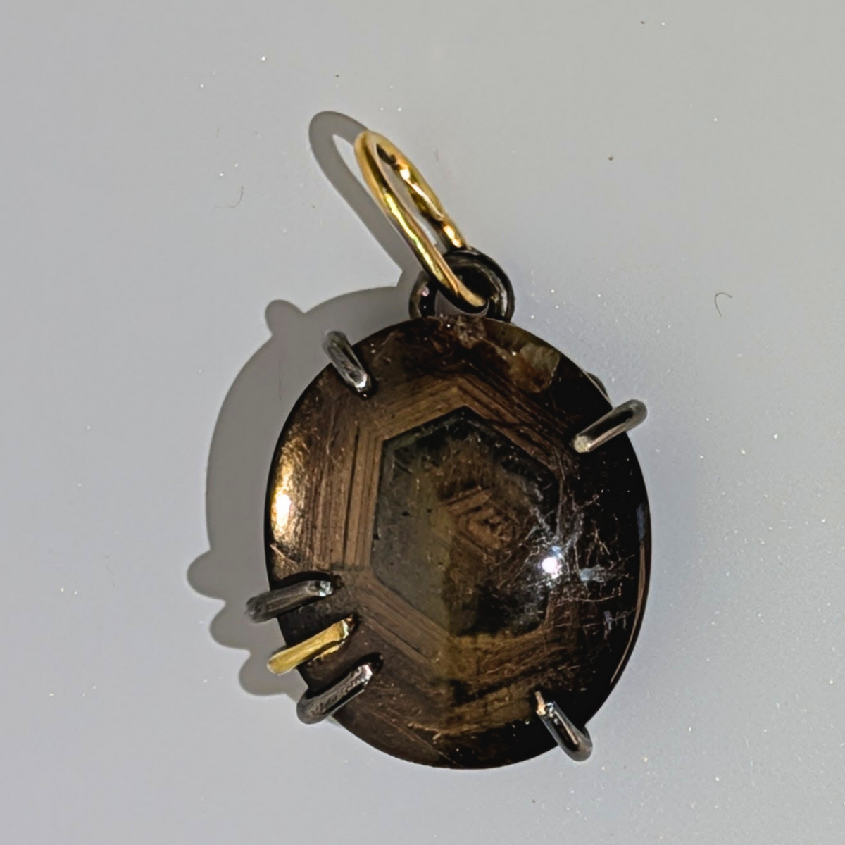 A handmade circular pendant featuring a dark brown textured center with a gold hexagonal design, held in silver prongs with gold and silver wire accents, on a light gray background.
