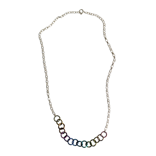 A silver chain necklace featuring a curved row of interlocking rings at the bottom, each with a different iridescent color finish, set against a plain white background.